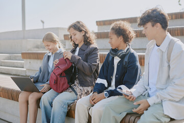 Fototapeta premium Biracial schoolkids sitting on wooden bench and resting before lessons