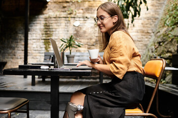 A young woman sits in a cozy cafe, sipping her coffee and focused on her laptop, showcasing her...