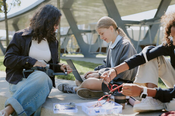 Teen girls helping their friend with making drone during warm day in park