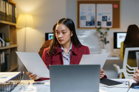Focused asian businesswoman works late in a dimly lit office, comparing documents on her laptop with determination and ambition