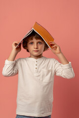 in a photo studio on a pink background stands a smiling schoolboy in a white sweater holding a book over his head preparing for school