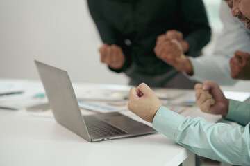 Business colleagues celebrating successful project outcome, raising fists in front of a laptop with charts, showing teamwork and achievement in office environment