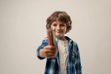 in photo studio on a white background stands a smiling schoolboy in a white sweater and a blue checkered shirt with colored pencils love for drawing