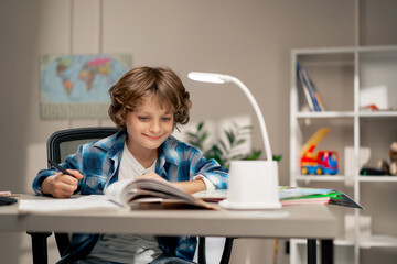 in the children's room a schoolboy in white sweater and a blue checkered shirt at a desk doing homework on his studies writing in a notebook