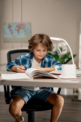 in the children's room a schoolboy in white sweater and a blue checkered shirt at a desk doing...