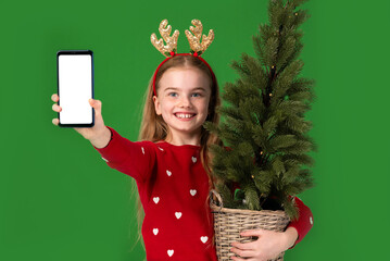 Cheerful Christmas child girl holding a smartphone with a blank white screen and holding a small Christmas tree. Christmas shopping and discount concept