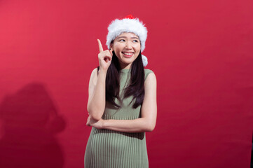 An Asian woman wearing a Santa hat, dressed in a sleeveless green dress, smiles brightly while raising her index finger, as if making a point, with a red background behind her