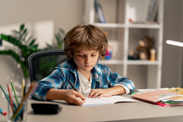 in the children's room a schoolboy in white sweater and a blue checkered shirt at a desk doing homework on his studies writing in a notebook