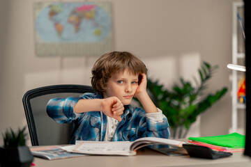 in the children's room a schoolboy in a white sweater and a blue checkered shirt at a desk doing homework on his studies writing difficult task children's suffering