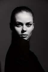 Confident young woman in black attire with a serious expression, posing against a dark background. Black and white portrait showcasing strength and elegance.