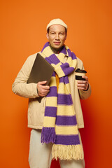 A tattooed young man confidently holds a coffee cup and laptop in a bright setting.