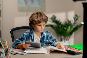 in the children's room a schoolboy in a white sweater and a blue checkered shirt at a desk does homework reads assignments on tablet modern education