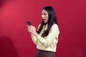 Fototapeta premium Asian woman looking at her smartphone with a surprised expression, wearing a yellow sweater against a red background. Her wide eyes and open mouth suggest she received unexpected news
