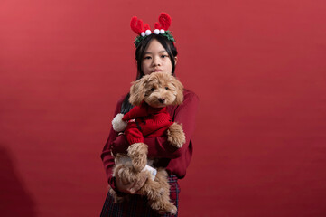 An Asian girl wearing reindeer antlers and a red sweater holds a small dog dressed in a red Christmas sweater against a red background. The scene exudes a festive, holiday spirit with cheerful