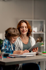 Fototapeta premium in the children's room a schoolboy in a white sweater and a blue checkered shirt at desk does homework mom explains the task