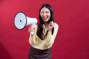Asian woman with long black hair holding a megaphone in front, speaking enthusiastically while wearing a yellow top and brown pants, set against a vibrant red background for a bold statement