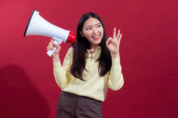 Asian woman with long black hair holding a megaphone in front, speaking enthusiastically while wearing a yellow top and brown pants, set against a vibrant red background for a bold statement