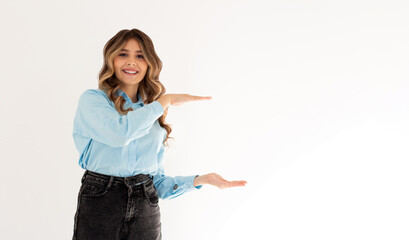 Young confident woman showing logo on empty space between arms. Smiling girl inviting or advertising promo deal, pointing hands right and looking at camera, white background.