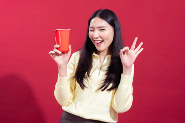 Asian woman smiling and holding a red plastic cup in her hand, wearing a yellow sweater against a red background. She looks cheerful and relaxed, with one arm crossed and the other holding cup