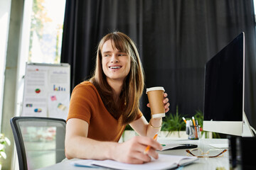 With a warm smile, a non binary individual sips coffee and engages in work tasks at a desk.