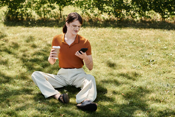 A non binary person sits on the grass, sipping coffee and using their phone.