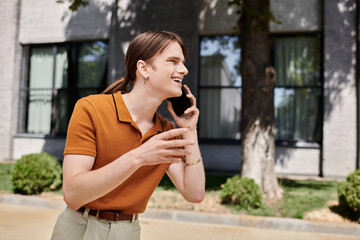 A young non binary person is happily talking on the phone, enjoying the sunny day outside.