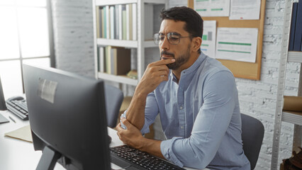 Young hispanic man working intently indoors at an office desk, deep in thought and focused on a...
