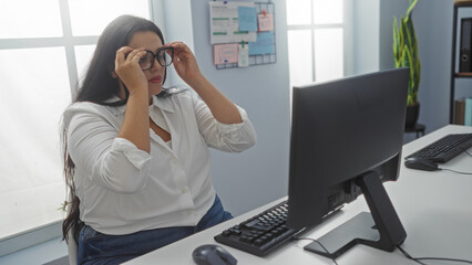 Woman adjusting glasses seated at a desk in an office, focusing on a computer screen with motivational notes on the wall behind her, creating a professional indoor workplace setting.