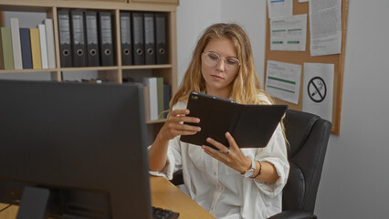 Young, blonde, woman working in an office, reading from a tablet at a desk, surrounded by shelves...
