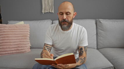 Handsome hispanic man with a beard sitting indoors on a couch in a living room writing in a journal