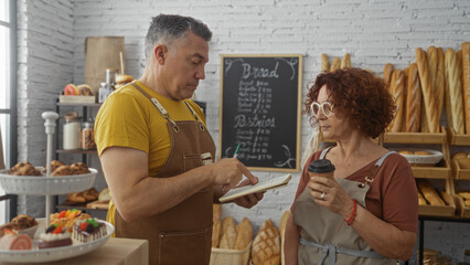 Man and woman bakers in a bakery discussing notes while she holds a coffee, with pastries and bread in the background.