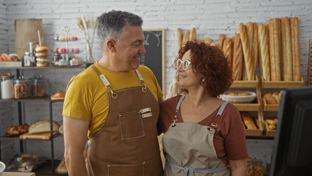 Man and woman bakers smiling together in an indoor bakery shop with shelves of bread and pastries behind them