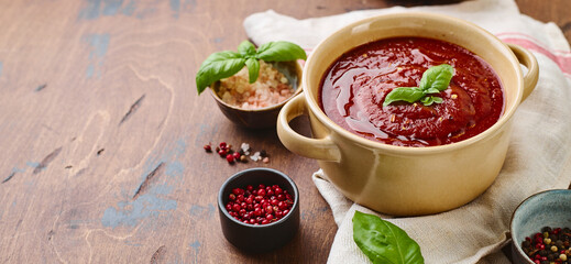 Tomato puree soup garnished with basil leaves and spices in ceramic bowl with toasted bread slices on wooden table