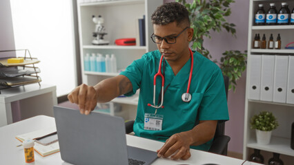 Hispanic male doctor wearing scrubs and stethoscope works on a laptop in a clinic office with...
