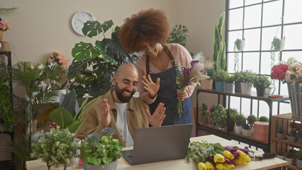 A woman and man, florists, laugh during a video call in a flower shop's interior surrounded by plants.