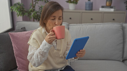 Mature woman sipping coffee while browsing a tablet on a cozy sofa indoors