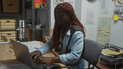 African american detective woman working on a laptop in a police station office filled with case files and a map.