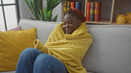 A content young african american woman relaxes on a couch, wrapped in a cozy yellow blanket, in a...