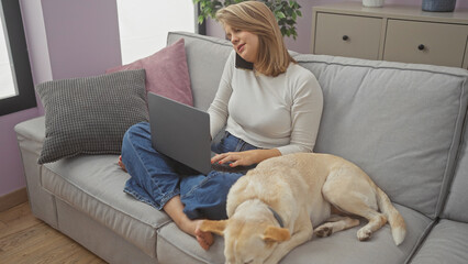 A woman multitasking at home, conversing on the phone while working on a laptop as her dog rests...