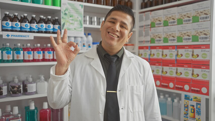Smiling young hispanic male pharmacist gesturing 'okay' inside a modern pharmacy