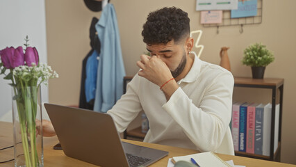 Stressed hispanic man using laptop in home office with flowers, bookshelf and casual wear.