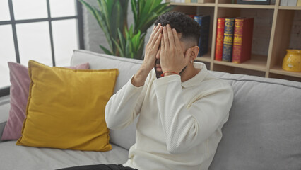 Hispanic man covering face with hands, expressing stress while sitting on a couch indoors, with colorful pillows and bookshelf background.