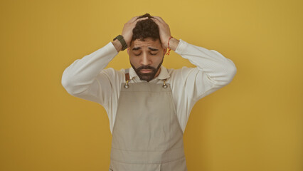 Stressed young man with beard in apron against yellow background