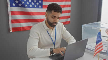 A young hispanic man with a beard works on a laptop in a room with an american flag and electoral...