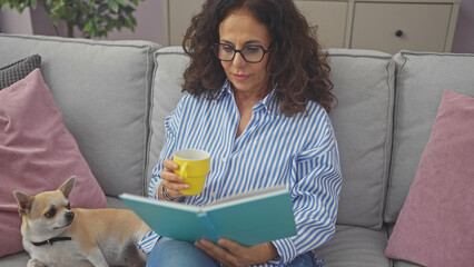 A middle-aged hispanic woman comfortably reads a book on a sofa with a chihuahua and a cup of...