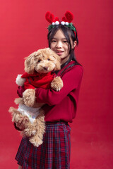 An Asian girl wearing reindeer antlers and a red sweater holds a small dog dressed in a red Christmas sweater against a red background. The scene exudes a festive, holiday spirit with cheerful