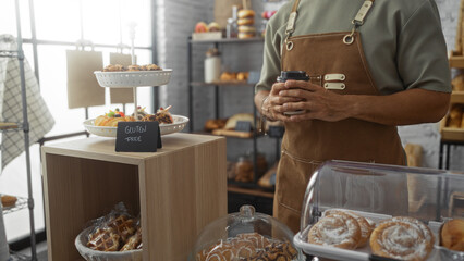 Man standing indoors in a bakery holding a coffee cup with gluten-free pastries on display