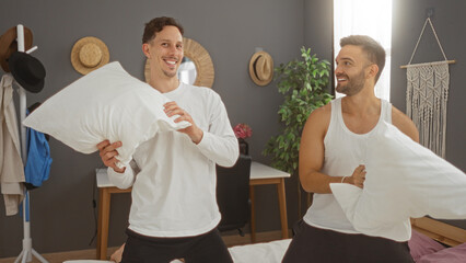 Fototapeta premium Men smiling while having a playful pillow fight on the bed in a cozy bedroom adorned with hats and wall decor, capturing a moment of love and joy in a home setting.