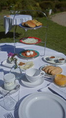 Sunny outdoor garden brunch with fresh pastries, cheese, nuts, tea, juice, yogurt, and flowers on a white tablecloth.