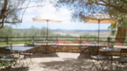 Fototapeta premium Blurred outdoor cafÃ© scene in mallorca with defocused tables and chairs under umbrellas amidst a scenic, sunny, mediterranean backdrop.